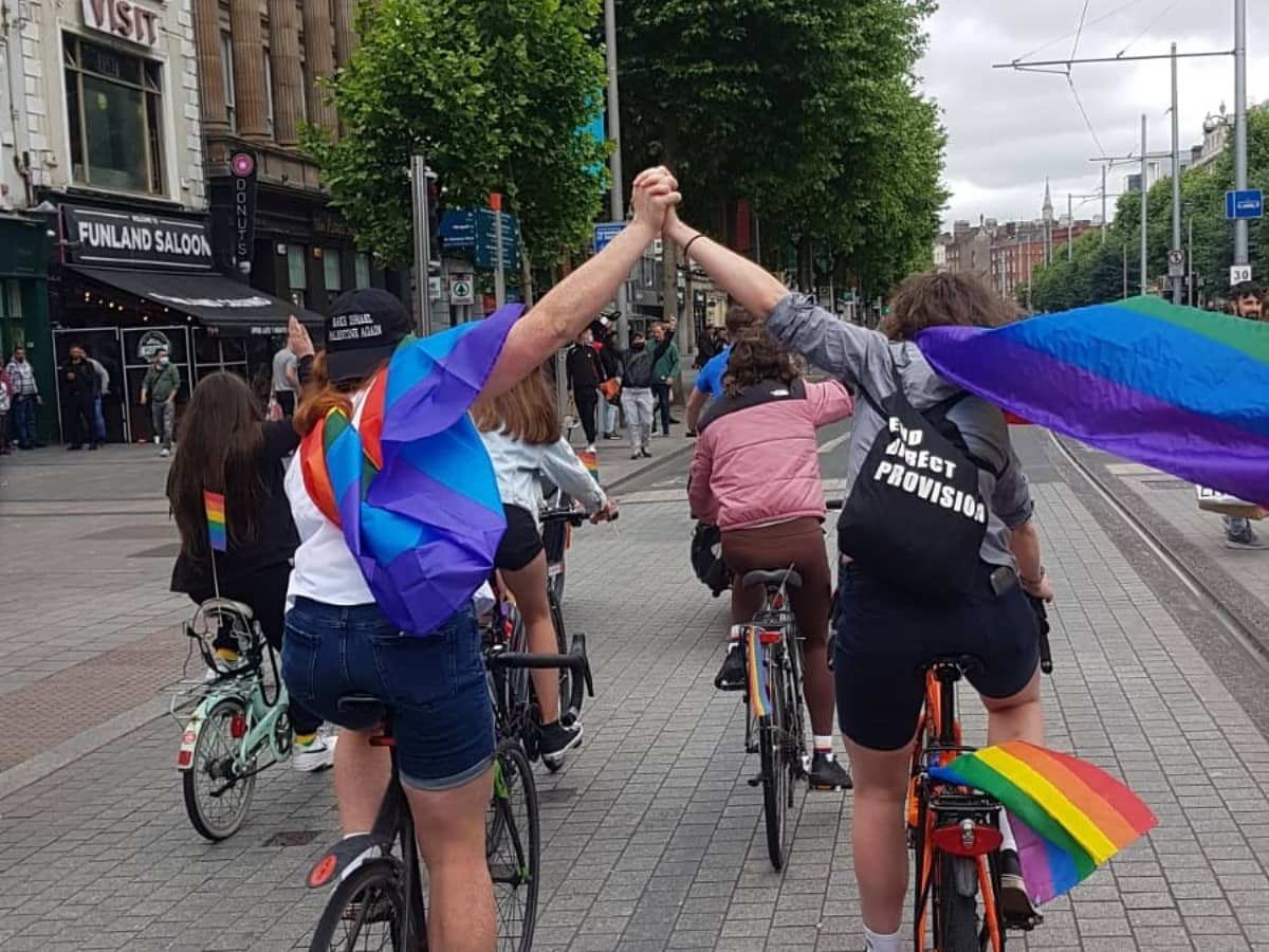 Image is a photo take during last year's PrideBikeBop. It shows two queer women cycling away from the camera as they travel north along O'Connell Street, Dublin 1. They're wearing pride flags on their backs (like capes, really) and are holding hands in the air. One of the women also has an "End Direct Provision" tote bag on her back, because end Direct Provision, dammit.