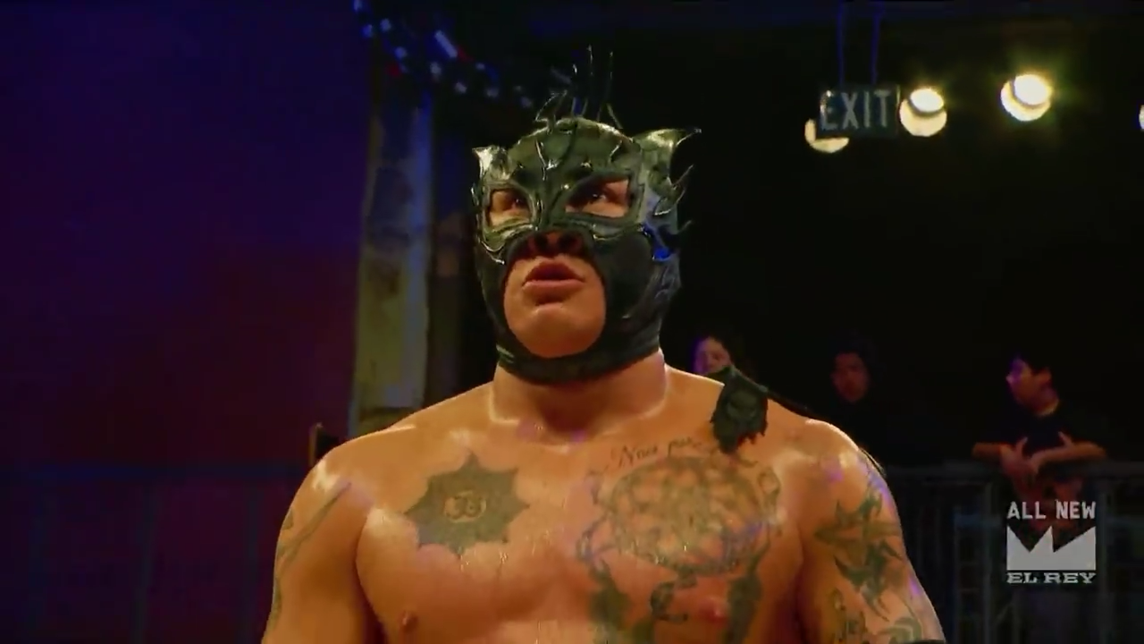 Screen cap from Lucha Underground. Fenix, a tan Latino luchador, is seen chest up. He is wearing a black mask vaguely resembling a bird with various tattoos on his chest. He is standing in front of the audience, looking dazed and unsure of where he is.