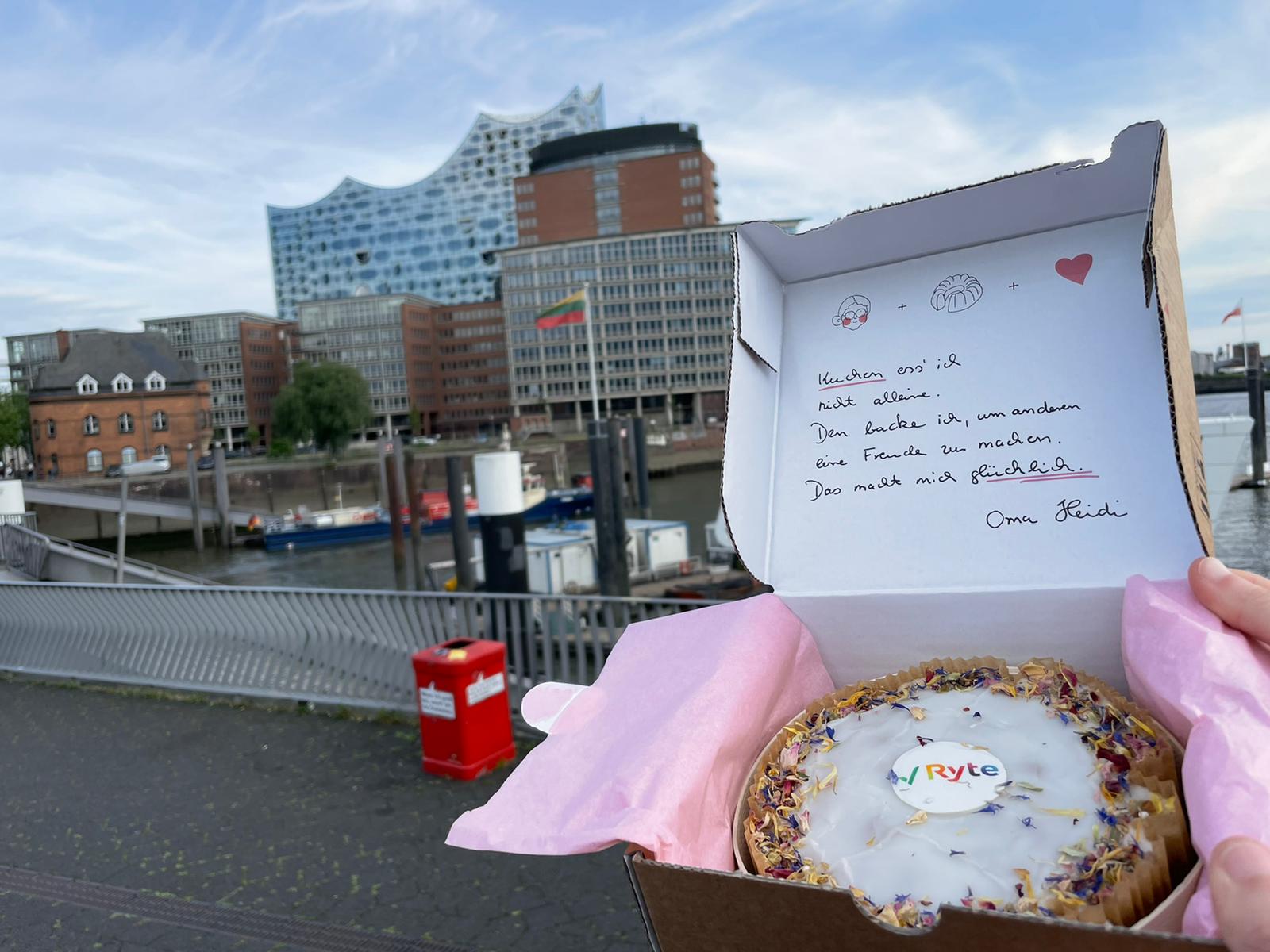 Kuchenbox mit Kuchen mit Ryte-Logo. Im Hintergrund sind Elbe und Elbphilharmonie zu sehen. Im Deckel der Box steht ein freundlicher Gruß von Oma Heidi.