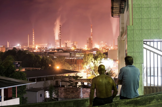 A imagem mostra dois homens de costas encontrados em uma mureta, eles estão observando a paisagem residencial e industrial da cidade de Ipatinga.