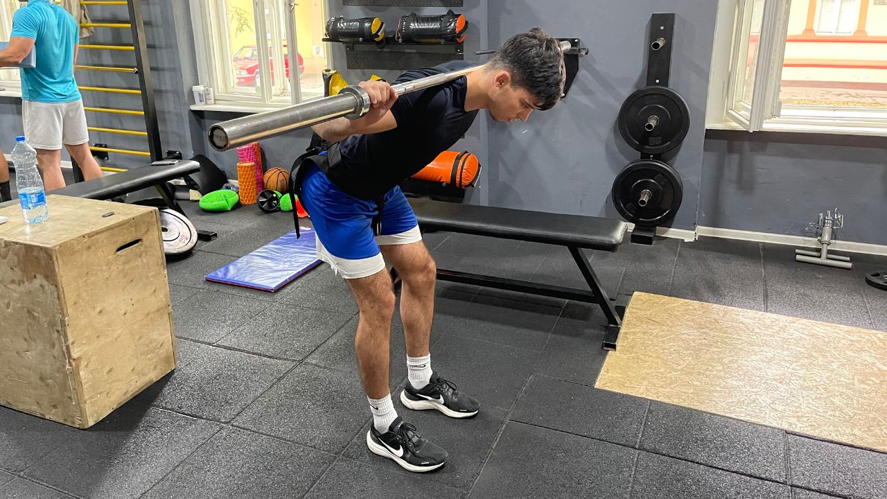 Vanja performs a barbell good morning exercise for the lower back in the commercial gym setup.