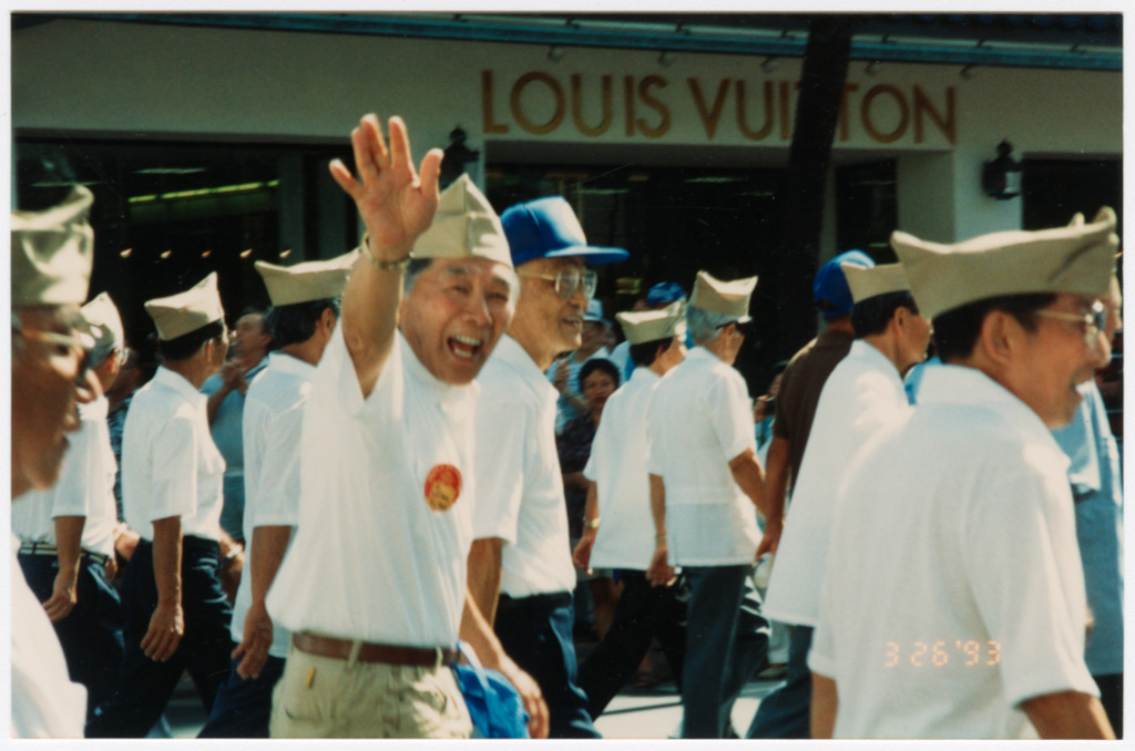 Veterans marching in a parade in Honolulu for the 50th anniversary reunion of the 442nd Regimental Combat Team. A veteran in the center of the photo is smiling and waving at the camera.