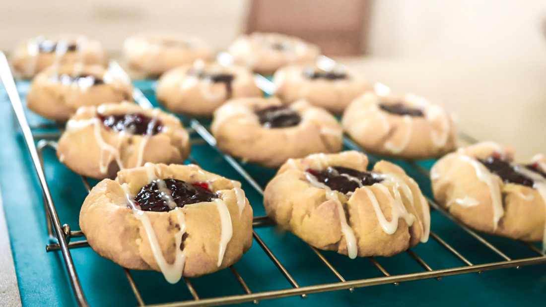 a tray of fruit-filled thumbprint cookies arranged on a cooling rack with a drizzle of white icing on top.