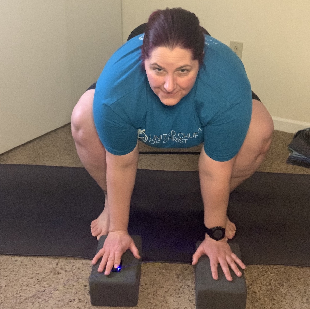 Nicole sitting on a folding chair that is placed on her purple yoga mat. She is bending over, her outer arms pressed against the inside of her knees. She is bending forward, reaching to gray yoga blocks placed in front of her.