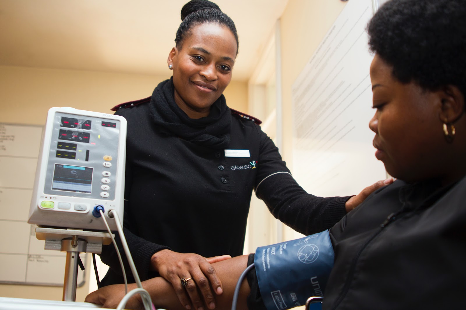 A smiling nurse who has applied a blood pressure cuff onto a seated woman’s arm looks into the camera as the machine takes a reading.