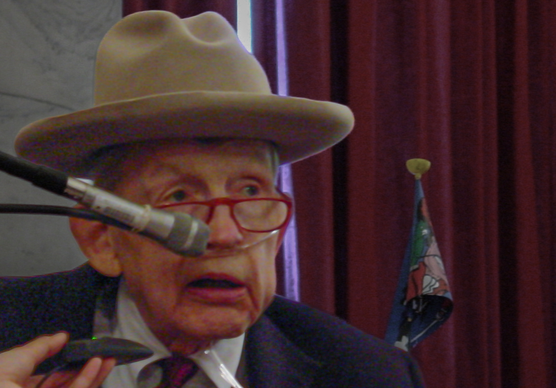 Man with cowboy hat, red glasses and oxygen tube to his nose speaks into a microphone 