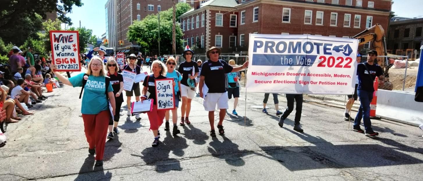 A photo of about 15 Voters Not Politicians volunteers walking in a parade. Two volunteers carry a large banner with the logo for Promote the Vote 2022 and text "Michiganders Deserve Accessible, Modern, Secure Elections!"