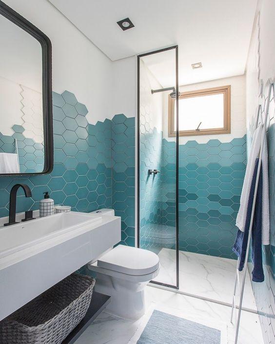 Banheiro com azulejo hexagonal meia parede por todo banheiro em degrade de tons de azul, piso porcelanato branco, bancada da pia branca com torneira preta e espelho e box de vidro com moldura preta