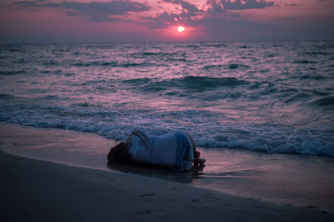A pink sunset over the sea. A woman is lying curled up on her side at the edge of the waves.