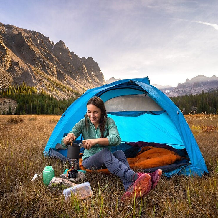 The chef behind Gritty  Gourmet, Karen Williams, cooks on a camp stove while sitting on the edge of her tent in a mountain valley