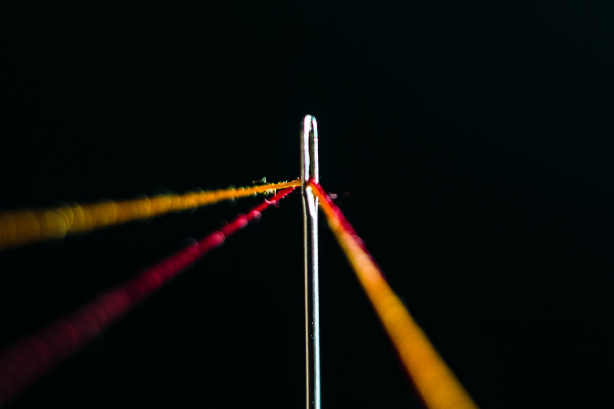 Close up shot of red and yellow thread in a needle against a black background.