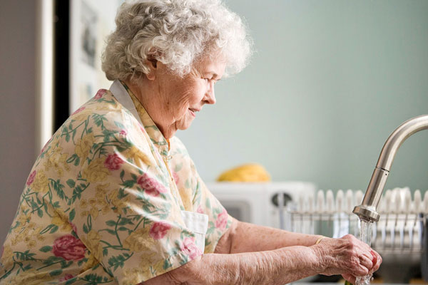 Woman with white hair and an apron on washing hands at a kitchen faucet.