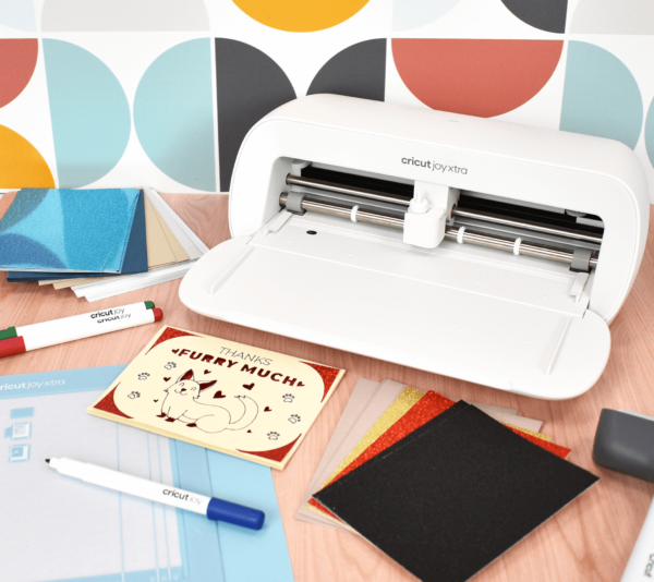 The image shows the Cricut Joy Xtra machine on a desk with crafting supplies.
