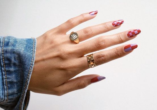 #DescriçãoDaImagem: mão com anéis dourados e unhas coloridas de rosa e lilás com estampas de quadrados grandes e pequenos. Foto: Pinterest.