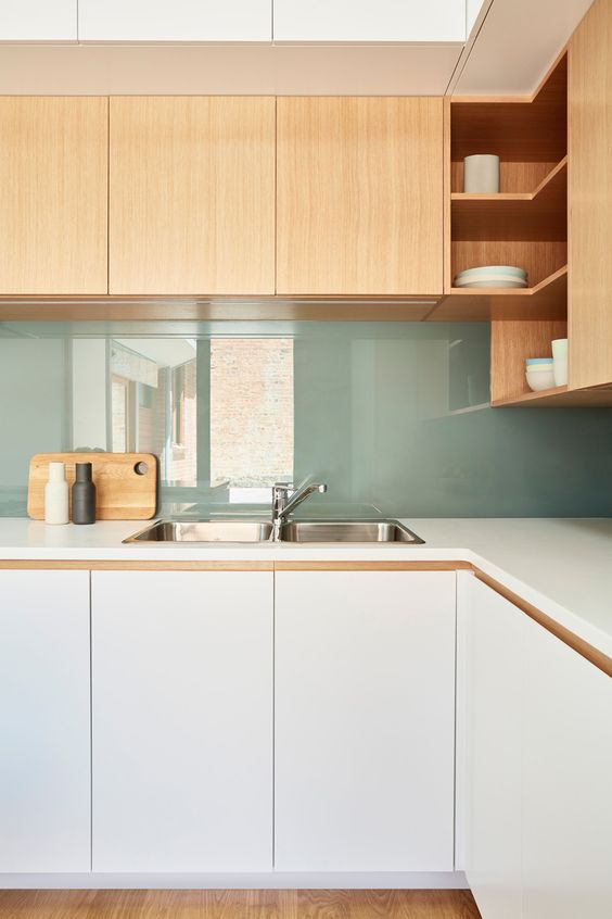 Cozinha com armários branco fosco, bancada branca, revestimento na cor verde claro e armários superiores de madeira de cor mel.