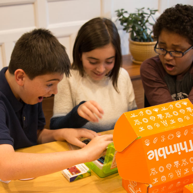 Three children are smiling at a table as they open a Thimble Creator Kit together.