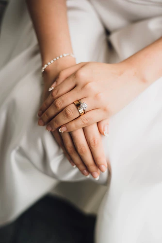 A woman’s hands over wedding gown with a ring and French nails displayed.

