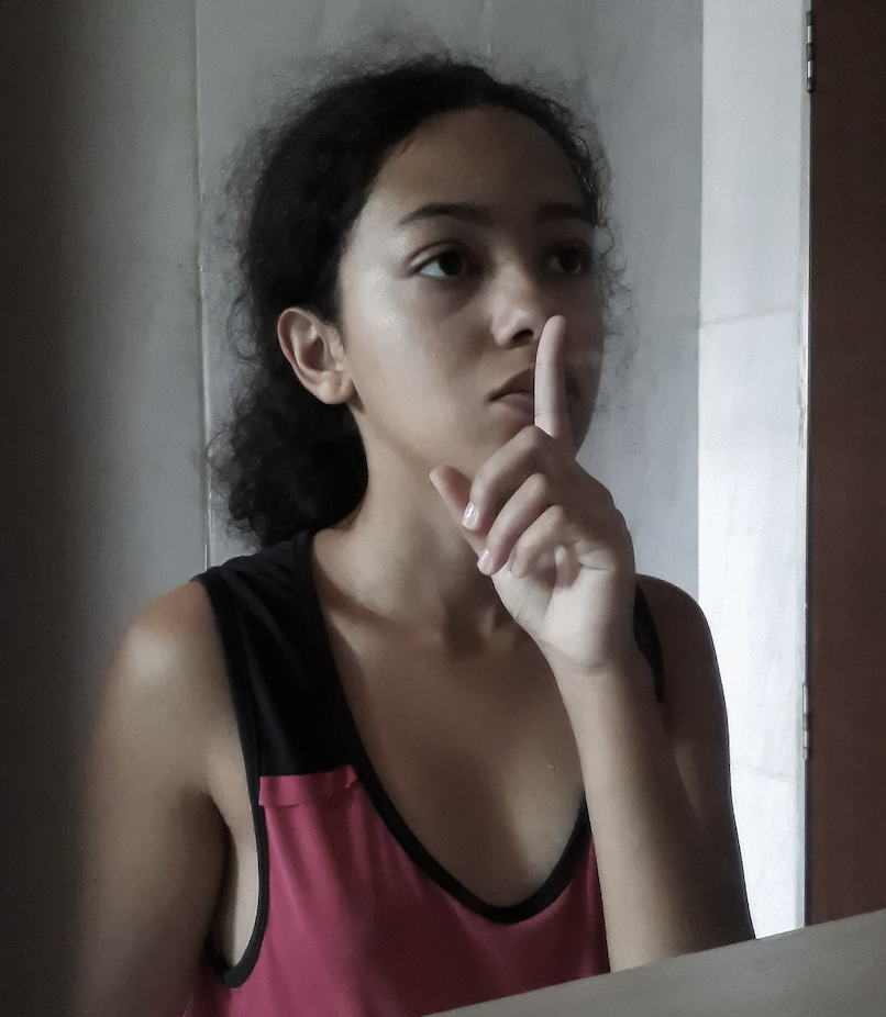 A young woman of colour in a pink tank top holds a hushing finger to her lips in the mirror.