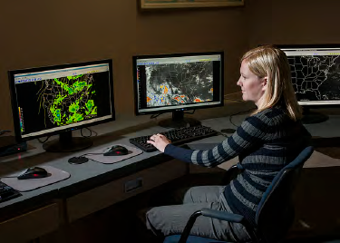 A female-presenting individual looking at 3 computer monitors in an office-like setting. Screens appear to show weather radar and U.S. map images.