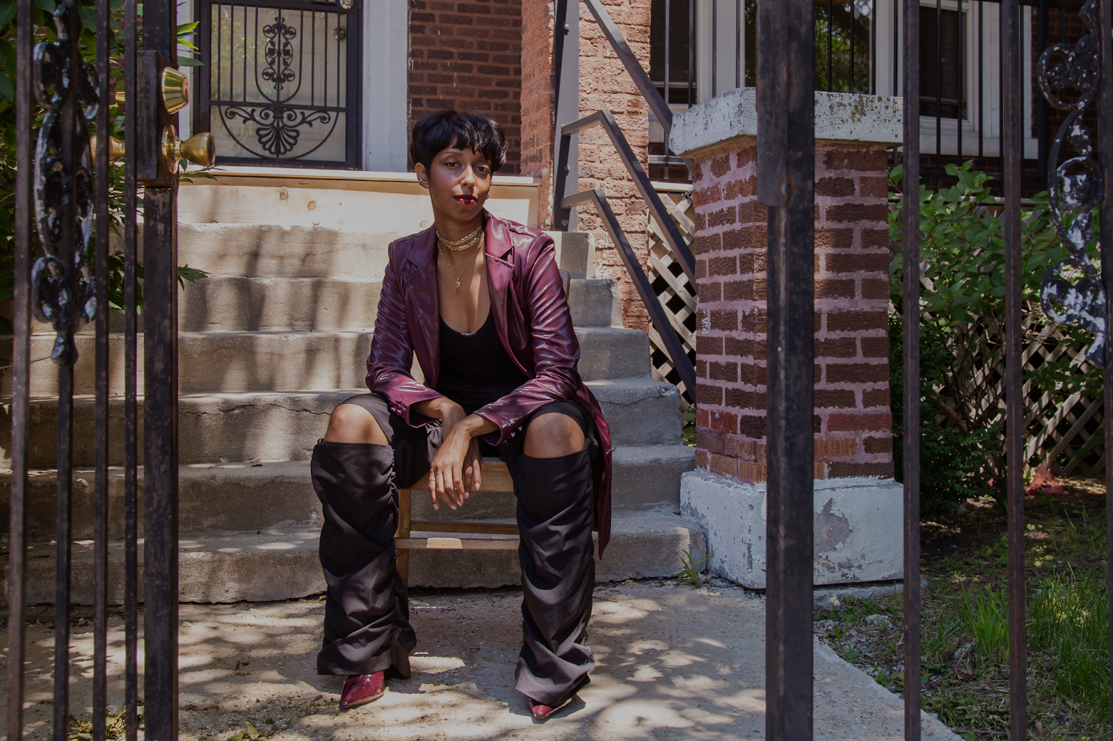 Image: Full image of Barédu sitting in front of a stoop on a wooden chair. Her arms are resting on her legs and she is wearing a long, burgundy jacket, a black top, black pants with knees cut out, and burgundy heeled boots. In front of her is a black gate, and behind her is a white door with a black gate. Image captured and edited by Joshua Johnson.