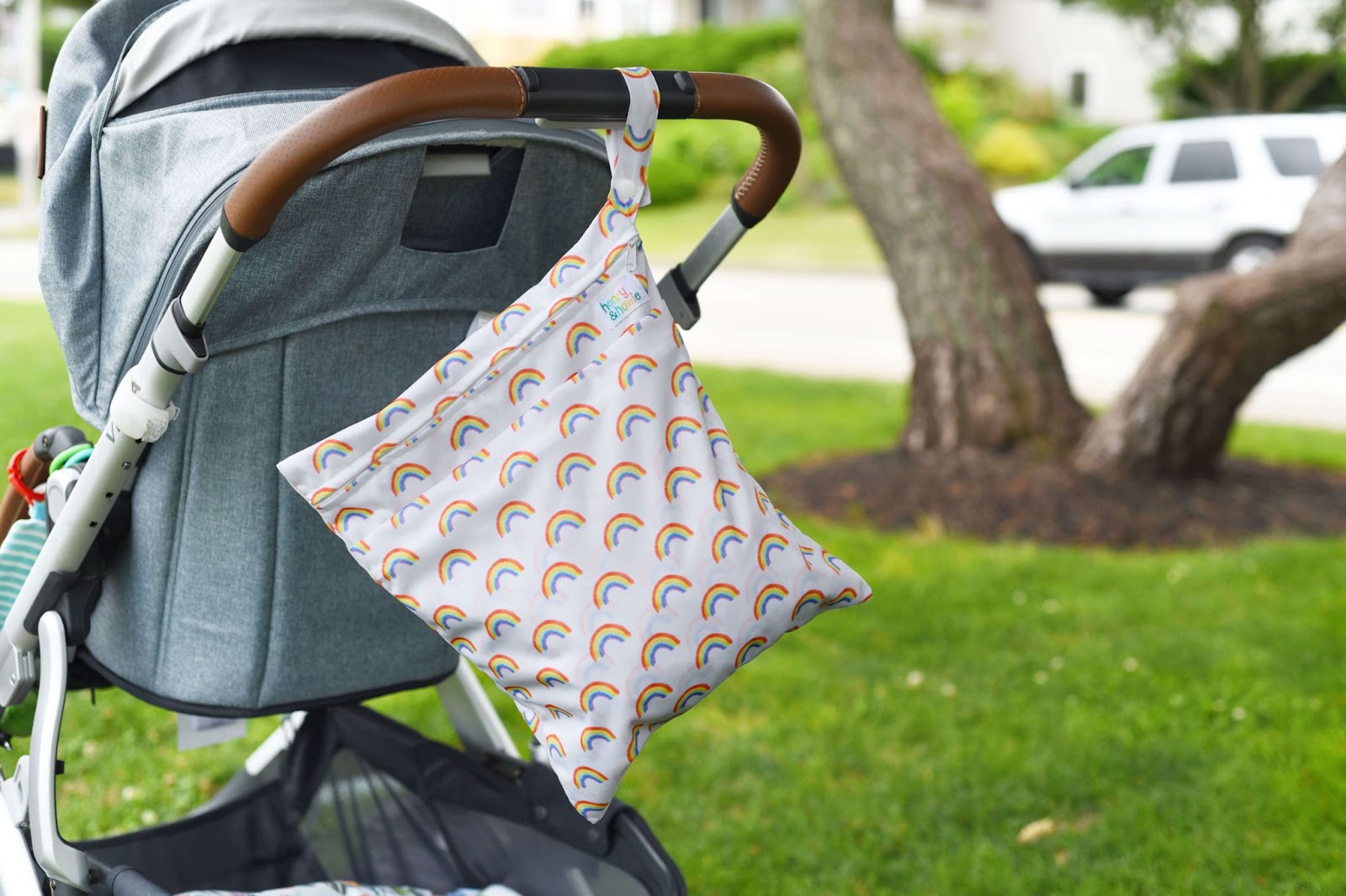 Making Cloth Diapering Cute Again With Henry and Howie–An image of a gray baby stroller on a patch of grass with a tree in the background. A Henry and Howie wet bag with a white background and colorful rainbow design is hanging off the handle of the stroller. 