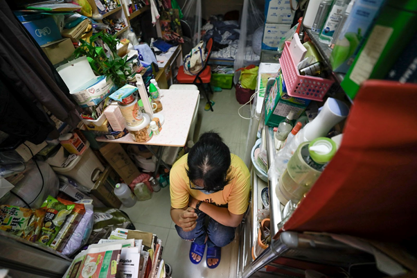 A photo of a person squatting in a tiny living space, packed on all sides with items, giving the impression that the person is in a closet, which is a home.