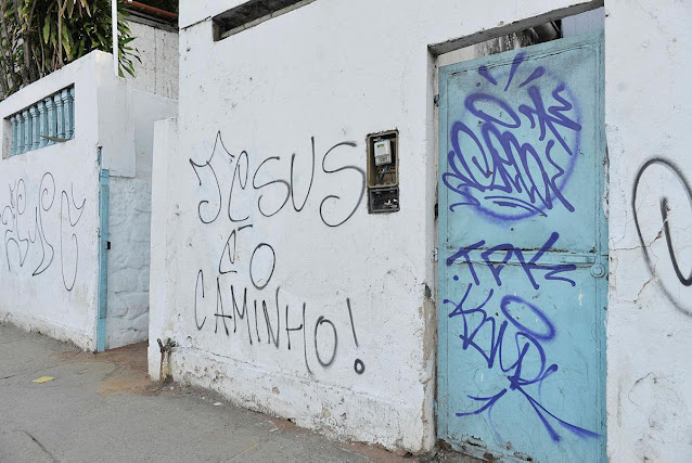 A fotografia mostra parte do muro do espaço de celebrações religiosas “Casa de Oxumarê” em Salvador (BA). No muro pintado de branco está pichado com tinta preta “Jesus é o Caminho”.