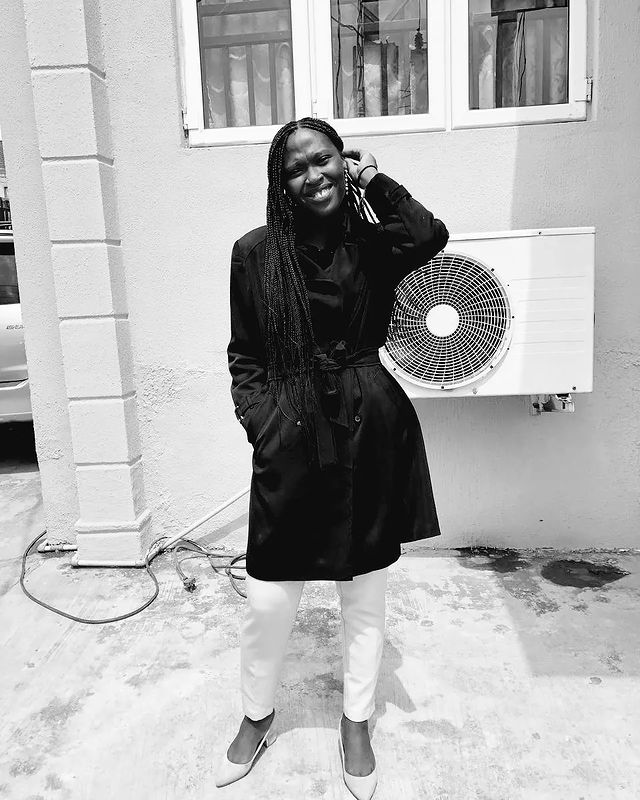 Black & white image: Nigerian woman looking towards the camera dressed in a black trench coat.