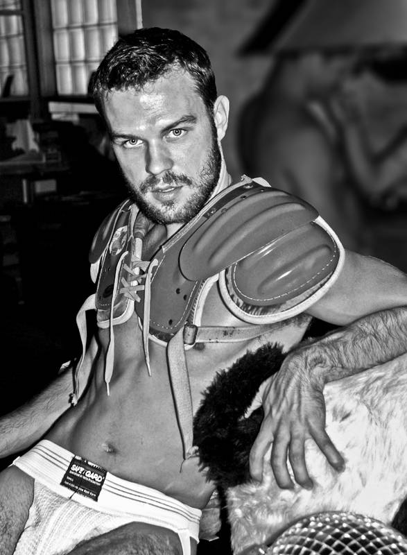 male model poses on a fur throw whirtless while wearing football shoulder pads and a white safe gard jockstrap