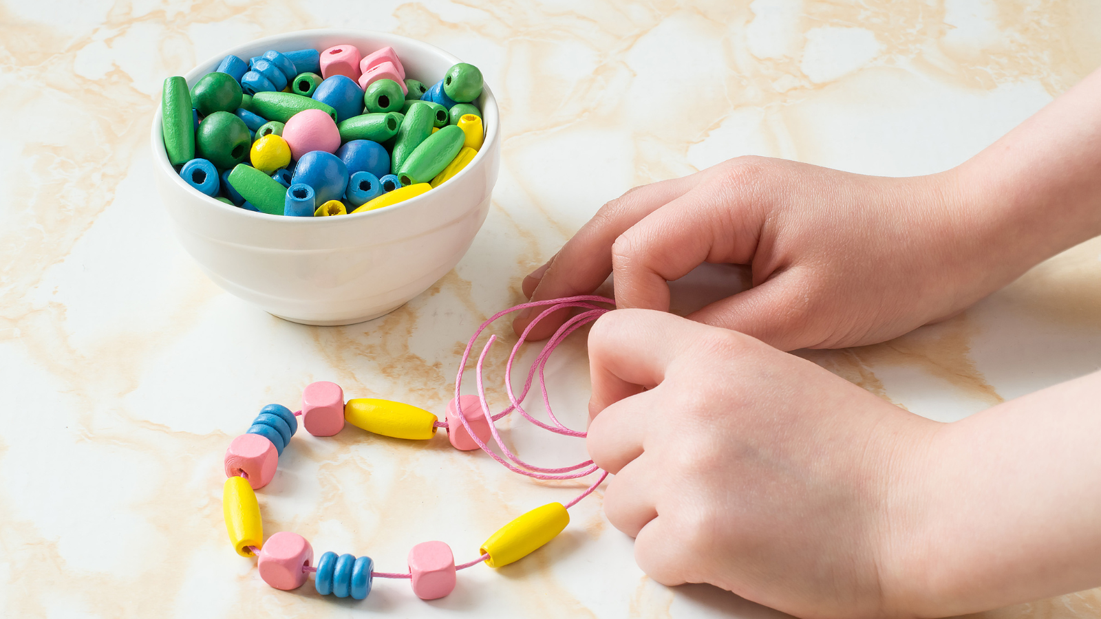 child stringing yellow, pink, and blue beads of different sizes on a pink string