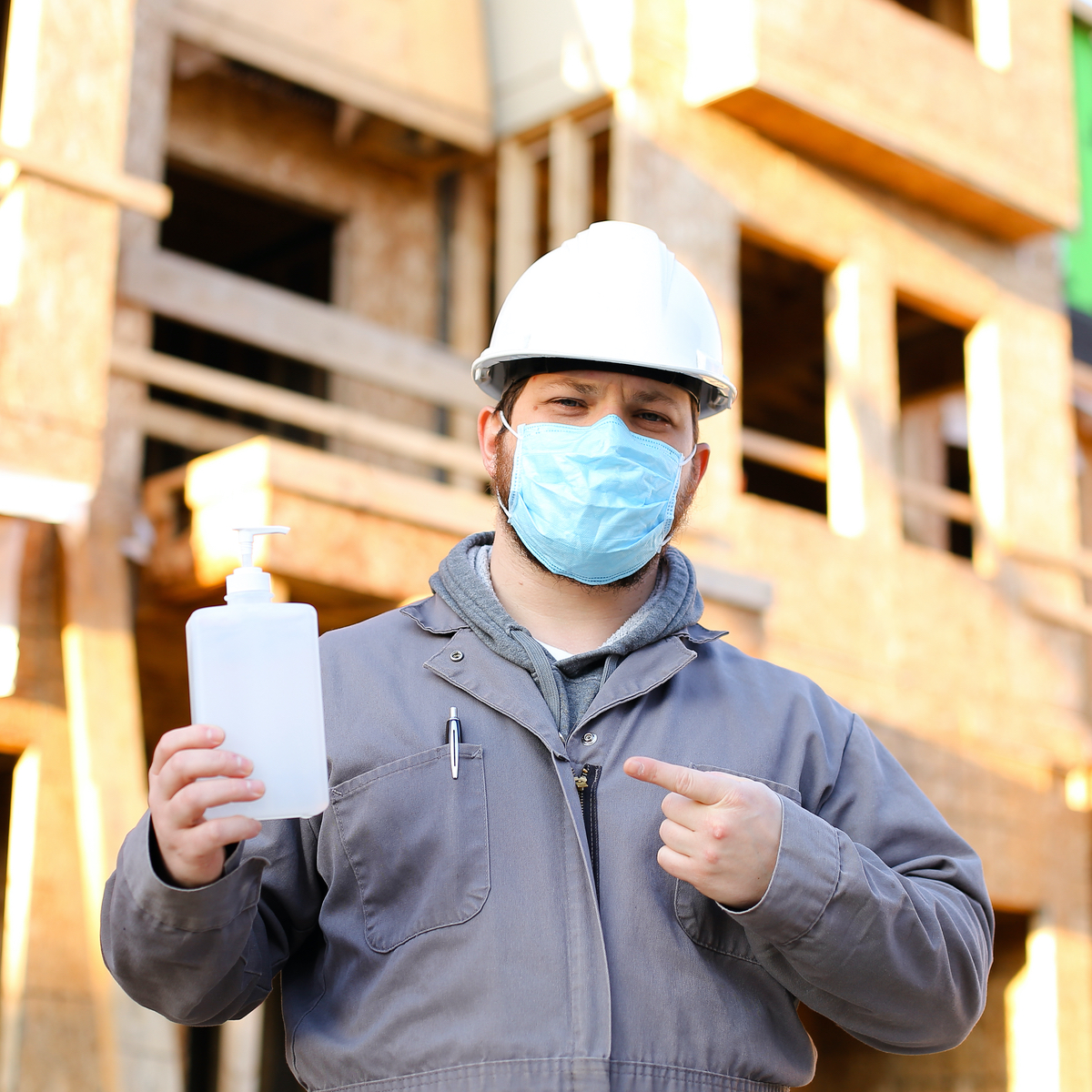 Masked construction foreman holding hand sanitizer in front of a home under construction
