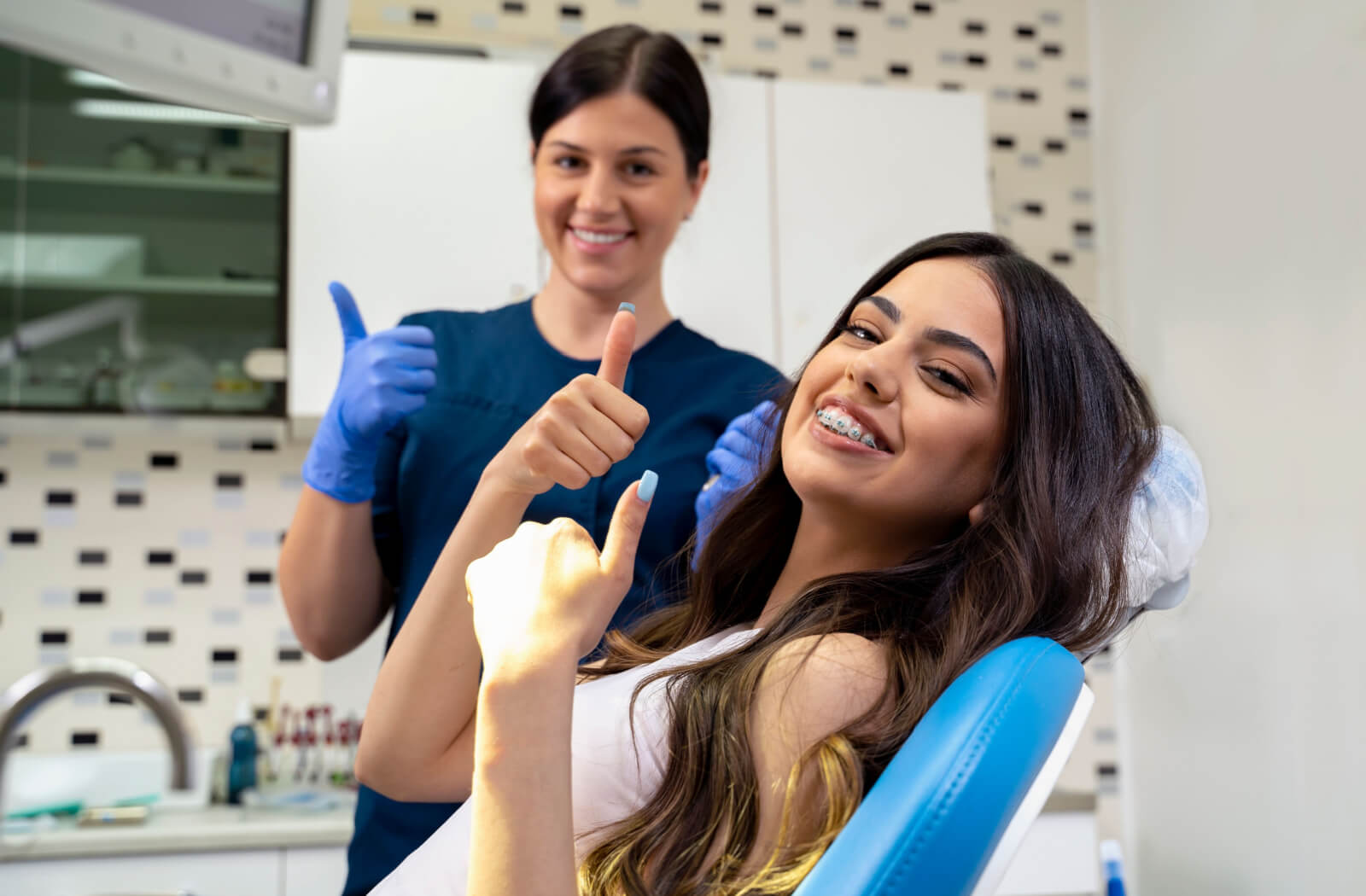 A young woman with braces smiling with her female dentist, giving a thumbs up while in the dental chair