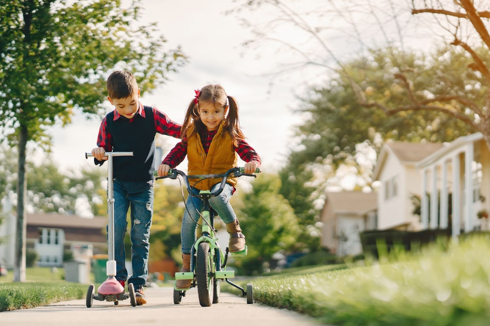 A boy and a girl riding a scooter and a bicycle respectively in a suburban environment