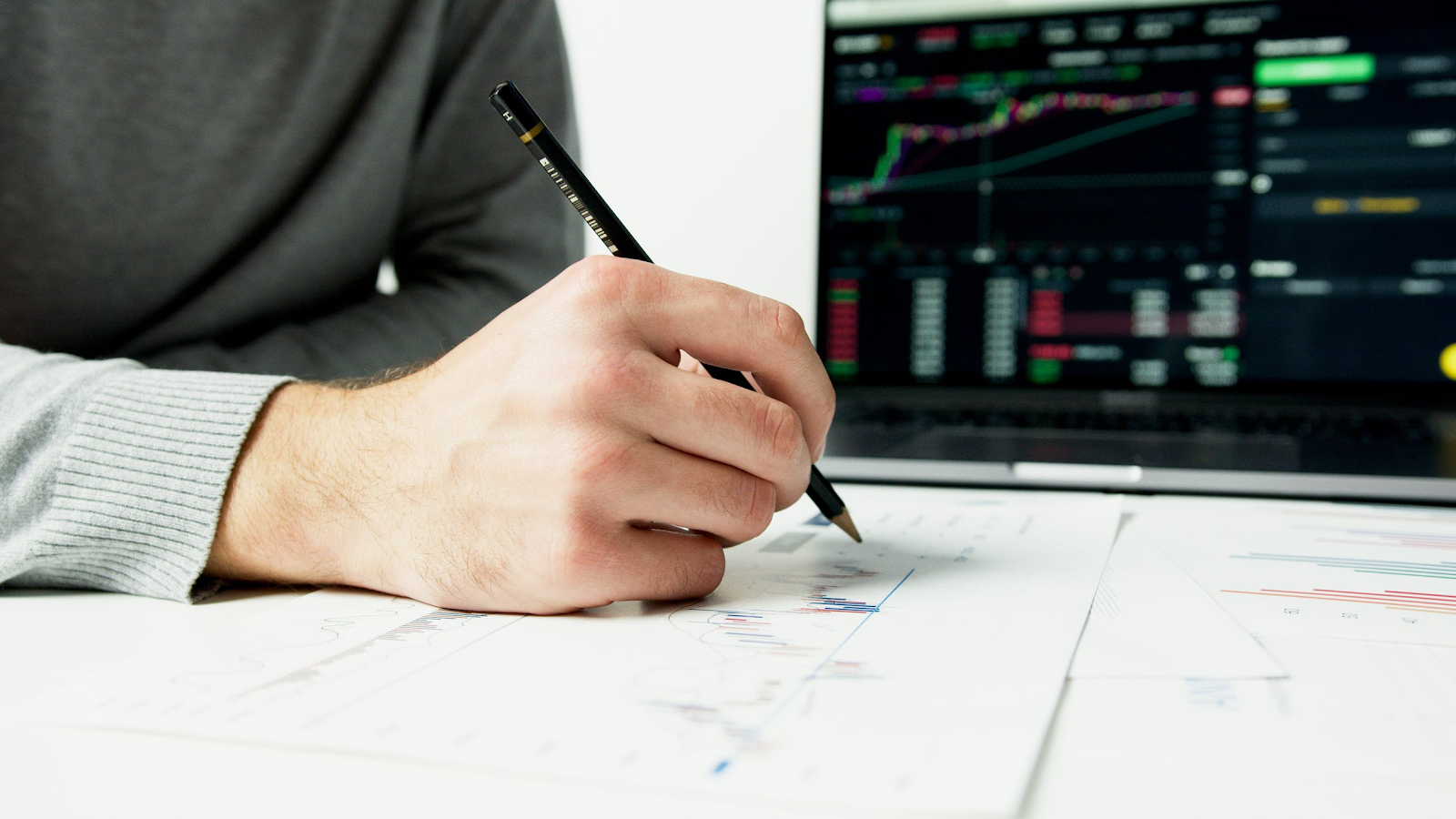 Person writing on white paper with a black monitor in the background.