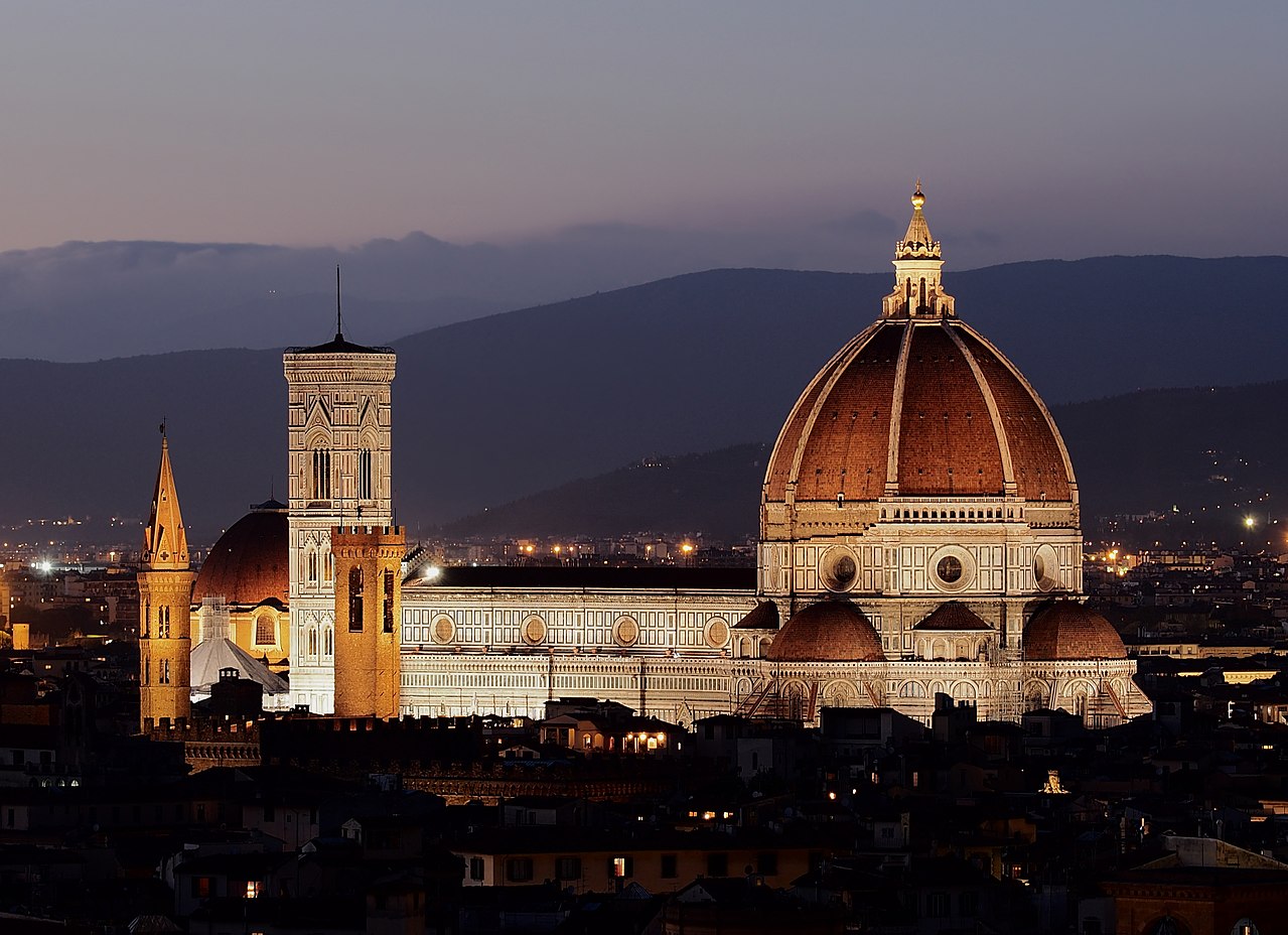 Photograph of a Renaissance era cathedral. This building has a tower on the lefthand side, and a very large dome on the right. Small mountains are in the background.
