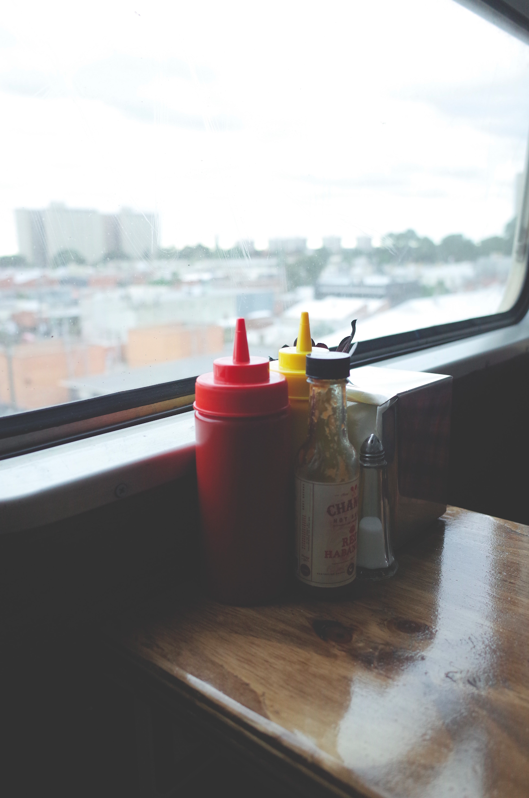 Close up shot of ketchup, mustard, salt, hot sauce, and a tissue dispenser near window.