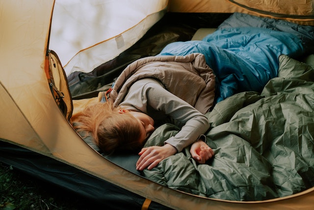 A woman sound asleep in a grey tent inside a blue sleeping bag. The tent is open, revealing a view of the outside. Kayak camping gear visible around her includes a sleeping pad.