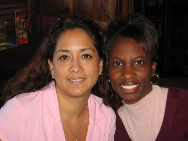Two female-presenting individuals, both people of color. They are leaning into each other and smiling. Only their heads and torsos are visible.  