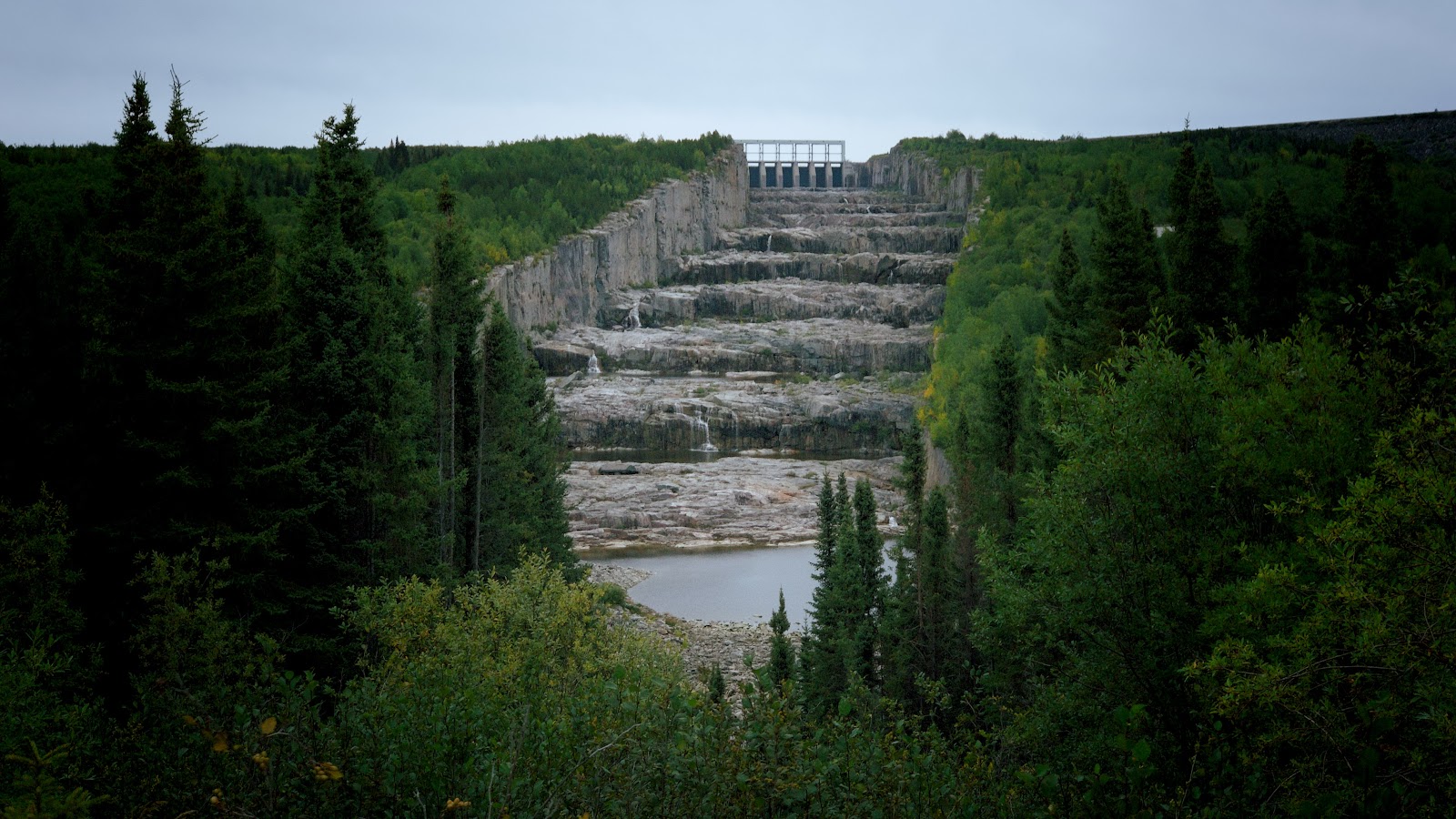 A picture of the Robert-Bourassa Dam with green grass and trees on each side and dam at the top, center of the image with stepped rocks in the middle. 