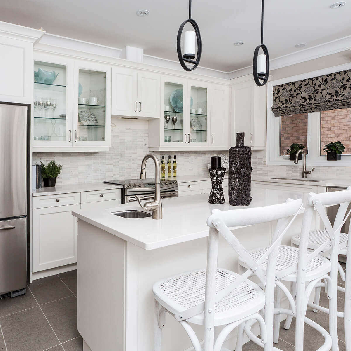 bright white kitchen fitted with white shaker cabinets. some of the shaker cabinets have glass doors, displaying dishware and decor inside. to match the cabinets, a bright white island with white countertops and matching chairs sits in the center of the room