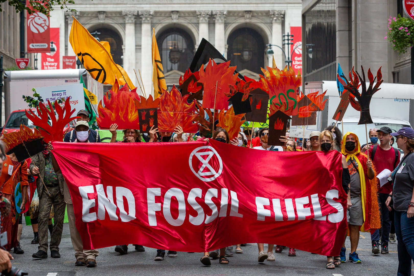 Rebels march through the street in a column with flags and fire imagery above and an end fossil fuels banner at the front.