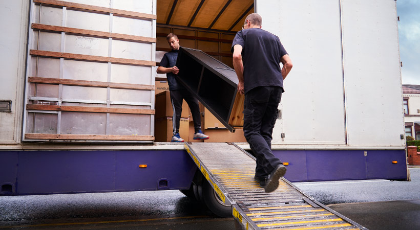 Full-service movers carrying a tall piece of wooden furniture up a ramp onto a moving truck.