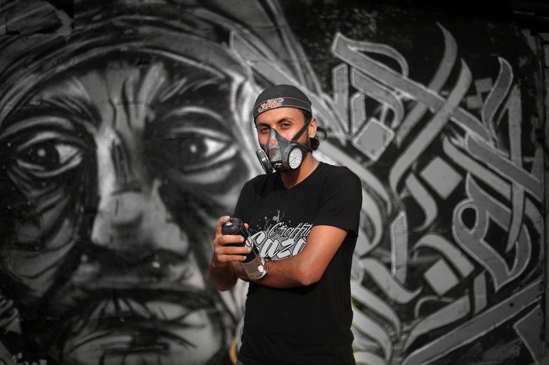 Young man wears baseball cap, gas mask and T-shirt reading Gaza graffiti while holding spray can in front of mural