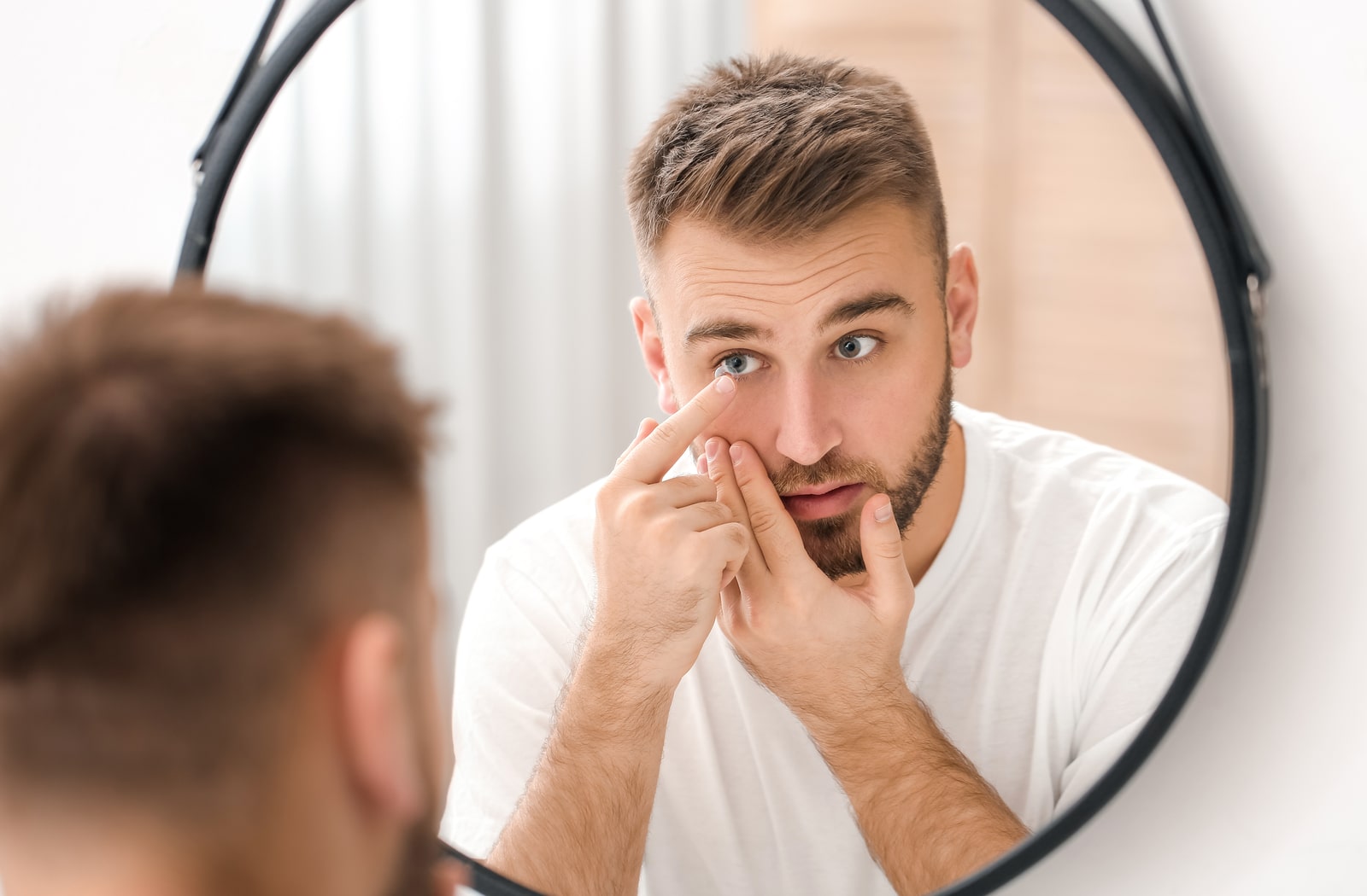 A man looking at himself in the mirror while he puts a contact lens in his eye