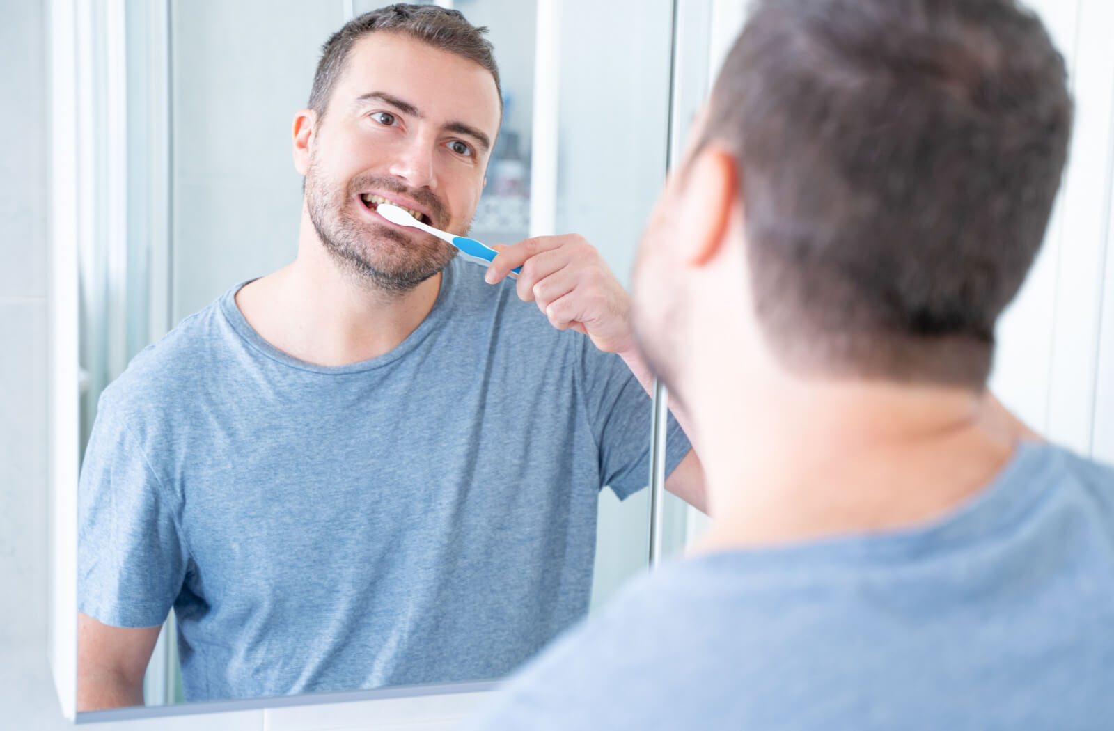 A man wearing a blue shirt with a toothbrush in his mouth smiling as he cleans his teeth.