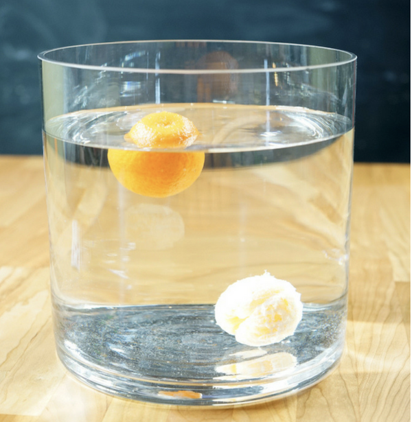 Glass of water with a whole mandarin orange floating in the water and a peeled mandarin orange sitting at the bottom of the glass of water.