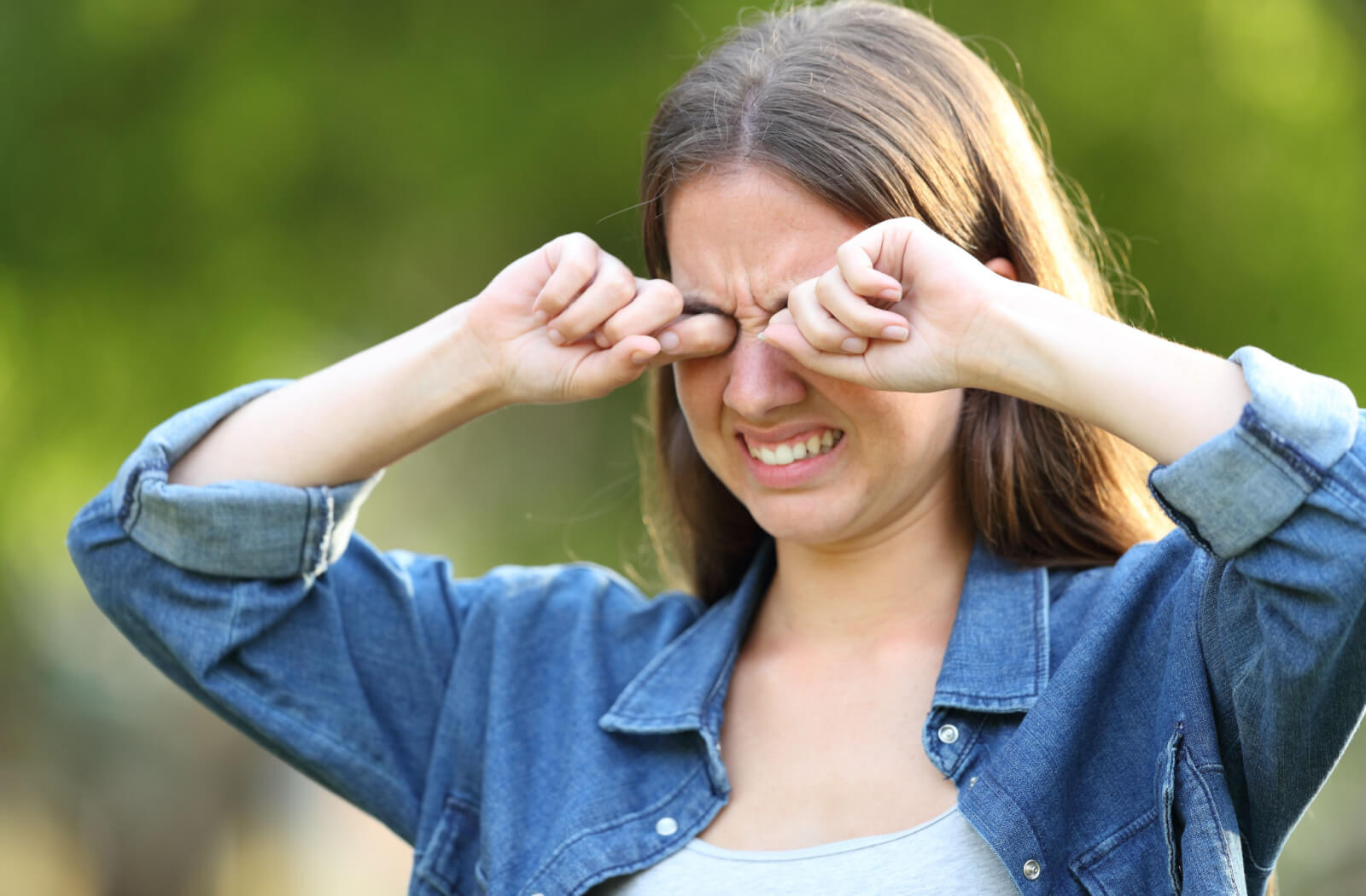 A young woman in a blue denim shirt is rubbing  her eyes, with an uneasy look on her face.