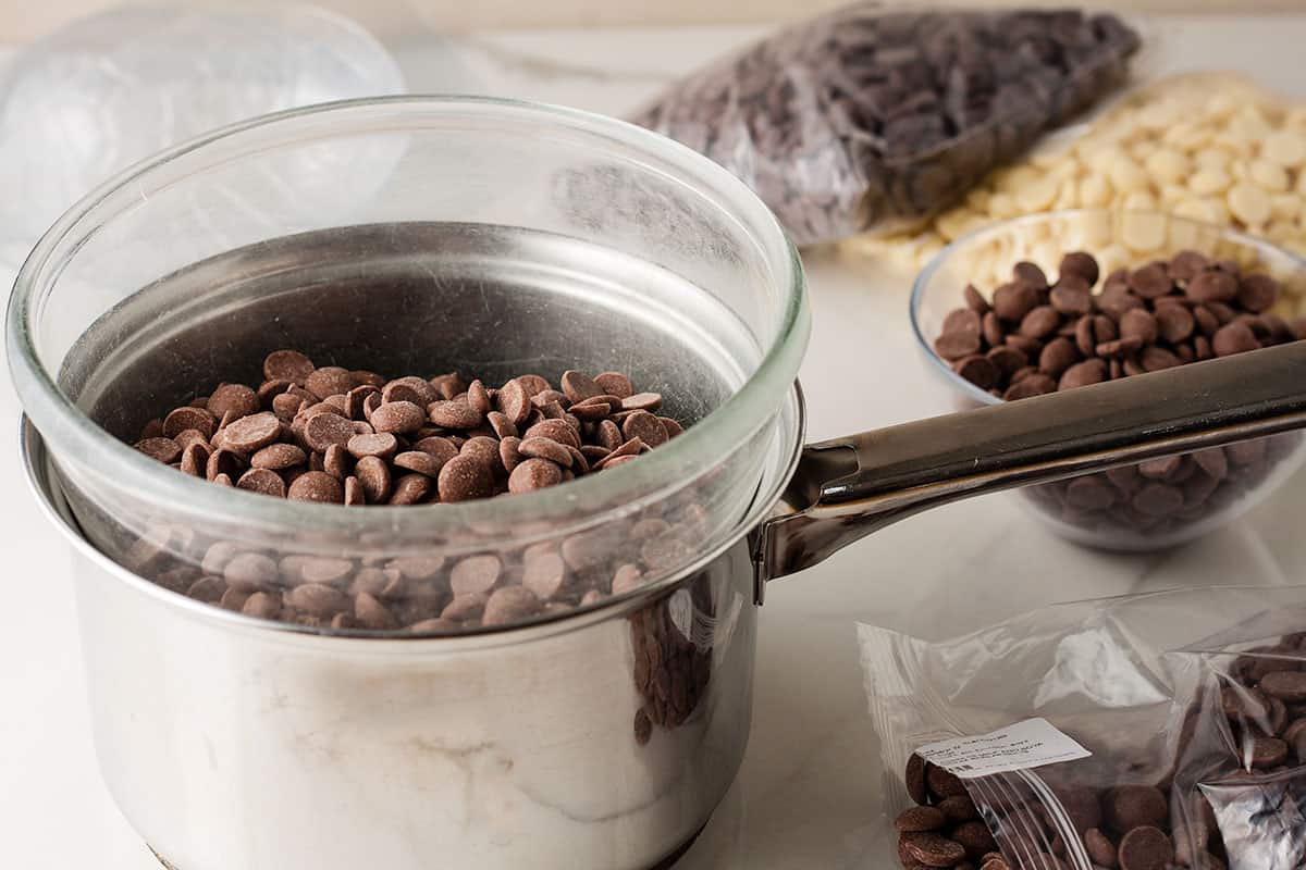 Bowl over saucepan filled with milk chocolate drops. Bags of chocolate behind.