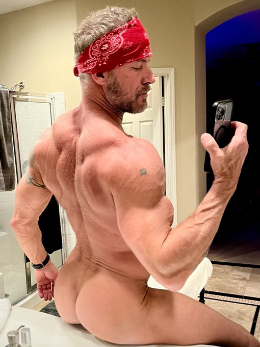 Greg Dixxon posing naked while sitting on the bathroom sink wearing nothing but a red bandana on his head