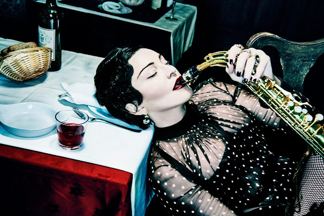 Fotografia da cantora Madonna, vestindo uma roupa preta de bolinhas brancas com um tecido transparente na parte dos ombros. Ela apoia a nuca em uma mesa enquanto toca um saxofone.